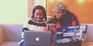 Image of two female-presenting Black individuals sitting next to each other, with one pointing to the other's laptop screen. The other person is smiling.